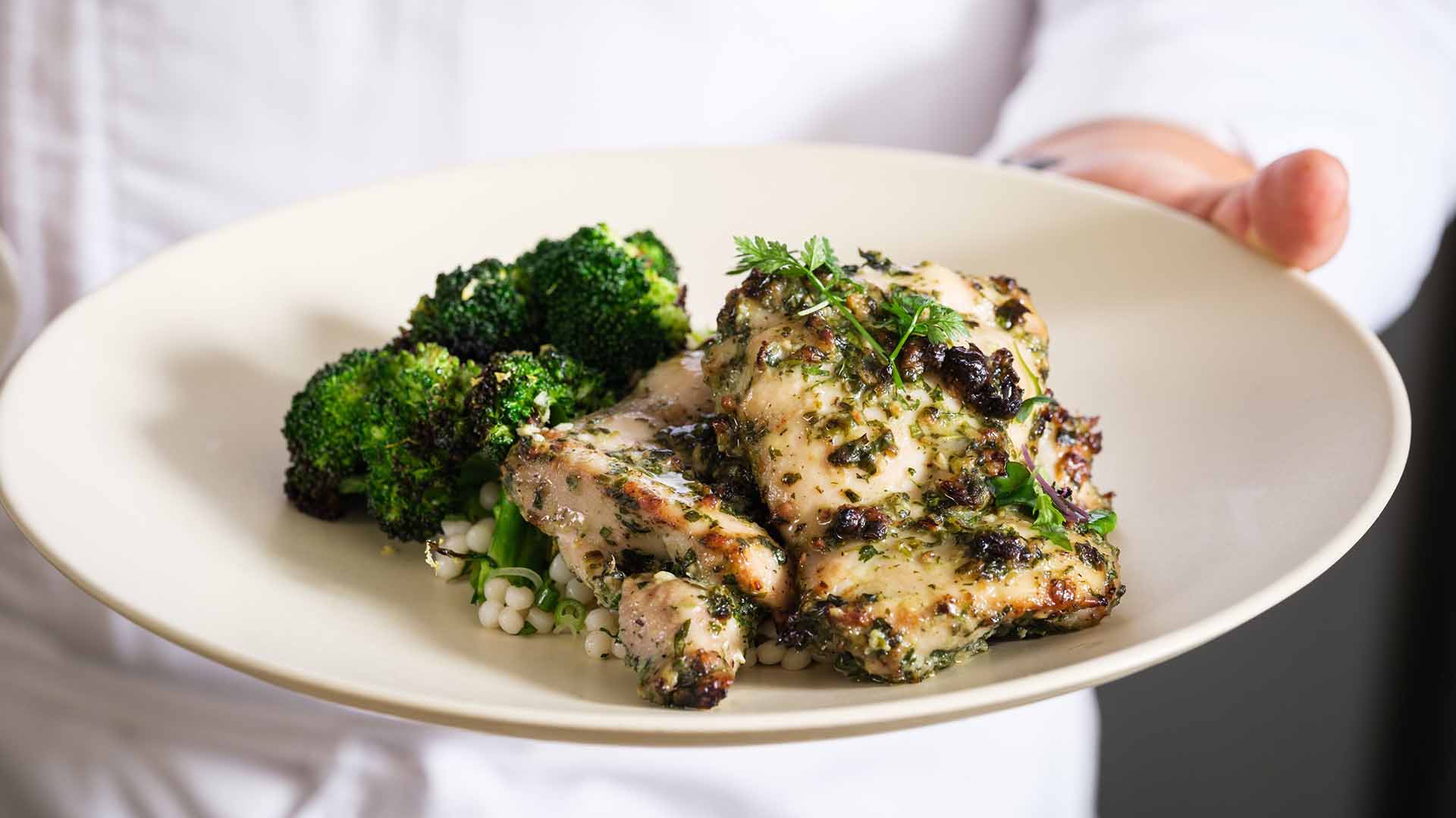 A person is holding a plate with grilled herb-seasoned chicken served alongside steamed broccoli and a small portion of grain.