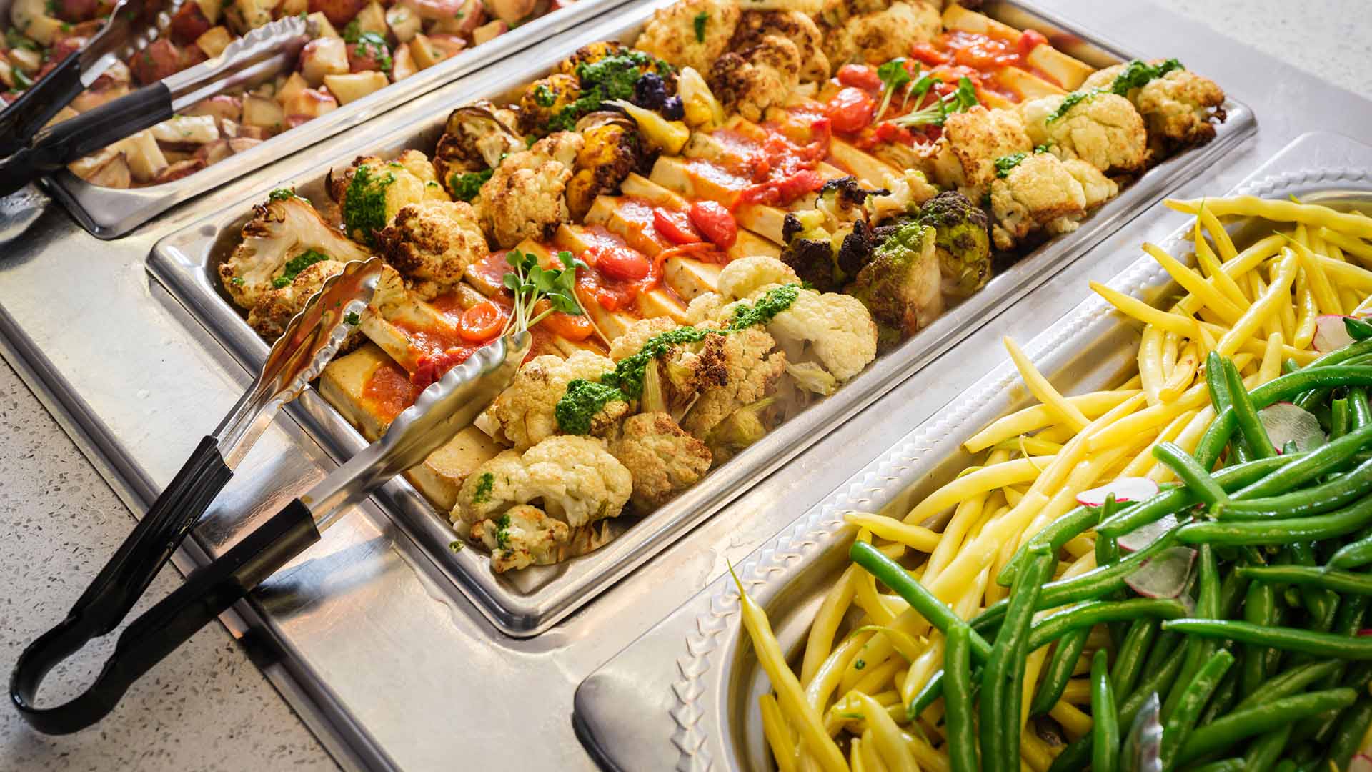 A buffet with trays of cooked vegetables, including green beans, yellow beans, cauliflower, grilled zucchini, and roasted potatoes, neatly arranged with serving tongs.